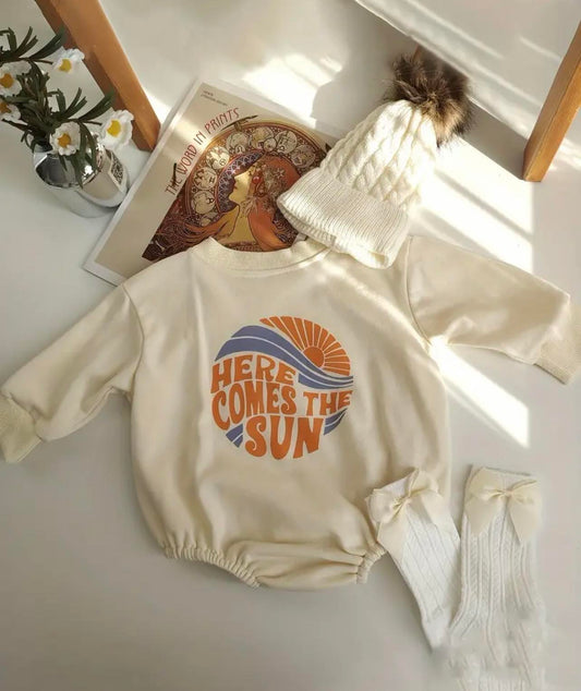 The Here Comes The Sun Bubble Sleeve Romper in 100% cotton, showcasing its oversized, relaxed fit with elasticized legs and a three popper fastening. The romper features a cheerful 'Here Comes the Sun' motto and is available in five sizes.